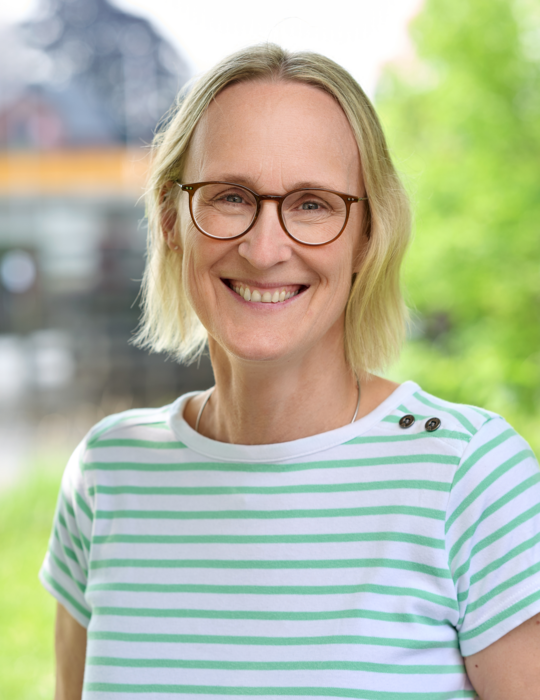 Foto von Birte Hüttmann. Sie lacht,  hat blonde , kinnlange Haare, trägt eine Brille und ein gestreiftes T-Shirt.